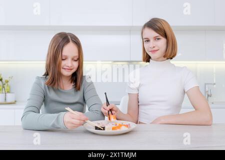 Zwei Mädchen essen Sushi in der Küche. Ein Mädchen hält Essstäbchen und das andere isst mit Gabel Stockfoto