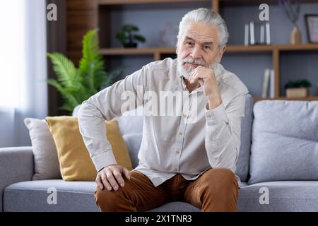 Ein älterer Mann mit stilvollem Bart und weißem Haar sitzt nachdenklich auf einer Couch, seine Hand auf dem Kinn und stellt einen ruhigen Moment in einem modernen Wohnzimmer dar. Stockfoto