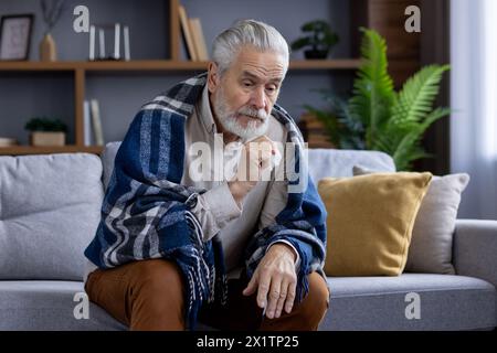 Seniorenmann mit grauen Haaren und Bart in eine gemütliche Decke gewickelt, sieht nachdenklich aus, während er auf einem Sofa in einem stilvollen Wohnzimmer sitzt. Stockfoto