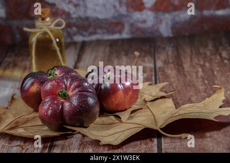 Gruppe von leckeren frischen Tomaten der blauen Sorte mit Wassertropfen auf trockenen Blättern neben Weizenohren mit einer Flasche Olivenöl im Hintergrund Stockfoto