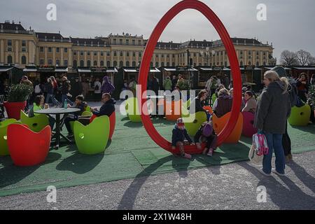 Lebhafter Ostermarkt mit Kindern und Erwachsenen, die auf bunten Apfelstühlen sitzen, mit einem großen rot symbolisierten Osterei im Hintergrund Schönbrunn Stockfoto