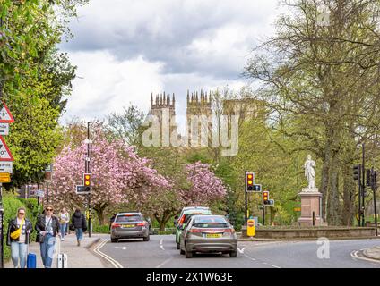 Eine Straßenkreuzung mit einem Bus an der Ampel. Die Türme des York Minster können über den Gipfeln der Bäume gesehen werden und ein wolkengefüllter Himmel ist darüber. Autos Stockfoto