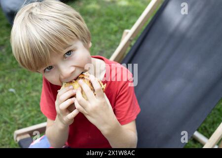 Positiver Junge beißt ein Sandwich während der Pause draußen im Park. Zeit für einen Snack auf einem Sommerspaziergang Stockfoto