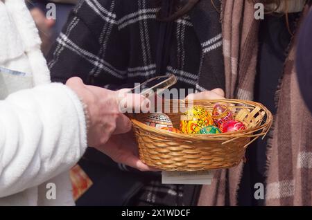 Mittelteil eines Kunden, Frau im Wintermantel, die Smartphone und Holzkorb hält und bemalte Eier kauft, traditionelle Osterdekoration im Na Stockfoto