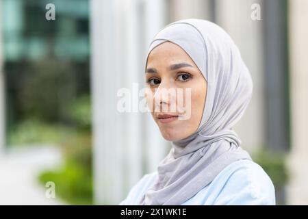 Porträt einer jungen muslimischen Frau, die einen Hijab trägt und selbstbewusst in die Kamera blickt, mit einem Hintergrund für Bürogebäude, der Professionalität und Eleganz vermittelt. Stockfoto