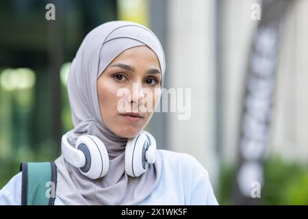 Eine nachdenkliche Frau mit Hijab und Kopfhörern, die ruhig aussieht und mit einem weichen Hintergrund in einer urbanen Umgebung aufgenommen wurde. Ein Bild, das modernen Lebensstil und Ruhe vermittelt. Stockfoto