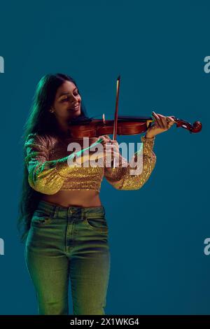 Künstlerische junge Frau in Glitzerspitze genießt es, Geige im Neonlicht vor verlaufendem Hintergrund zu spielen. Stockfoto