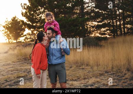 Familie genießt einen ruhigen Spaziergang in der Natur - Kind reitet glücklich auf den Schultern ihres Vaters - Mutter küsst ihn: Zuneigung und Zweisamkeit in der Abenddämmerung. Stockfoto