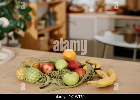 Verschiedene Früchte in der Einkaufstasche, die auf der Küchentheke oder auf dem Holztisch vor dem Innenraum mit grünen Hauspflanzen steht Stockfoto