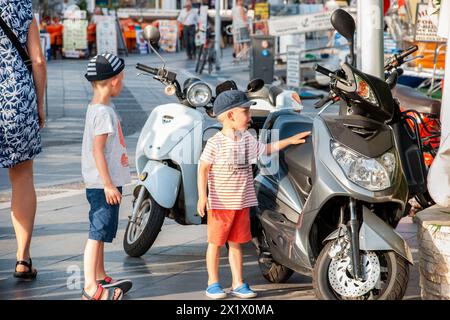 Zwei kleine Jungen in legeren Sommerkleidung bewundern die Motorroller, die auf der Straße im Resort geparkt sind Stockfoto