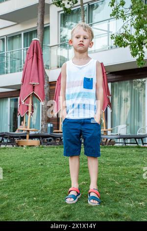 Trauriger Junge in Strandkleidung steht auf einem gepflegten Rasen vor dem Hintergrund eines modernen Strandhotels mit geschlossenen roten Sonnenschirmen und Liegestühlen Stockfoto