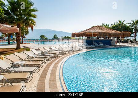 Kreisförmiger Swimmingpool, umgeben von Liegestühlen und einer Bar mit Strohdach, in einem Sommer-Wasserpark am frühen Morgen Stockfoto