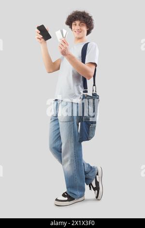 Junger Mann lächelnd und gutaussehend, der Handy und Kreditkarte in Jeans und T-Shirt mit Schultertasche zeigt, isoliert auf grauem Hintergrund Stockfoto