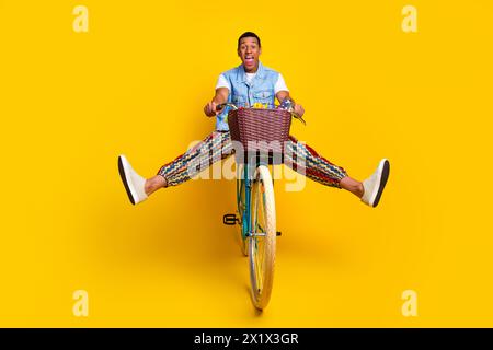 Foto in voller Länge des exzentrischen lustigen Guy gekleidete Jeans Weste Fahrt auf dem Zyklus erstaunt starrend isoliert auf gelbem Hintergrund Stockfoto