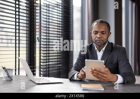 Fokussierter Unternehmer, der durch ein digitales Tablet scrollen kann, während er mit Jalousie am Fenster arbeitet. Aufmerksamer Finanzmanager, der Daten zu Unternehmensrechnungen in der Systemanwendung des Unternehmens eingibt. Stockfoto