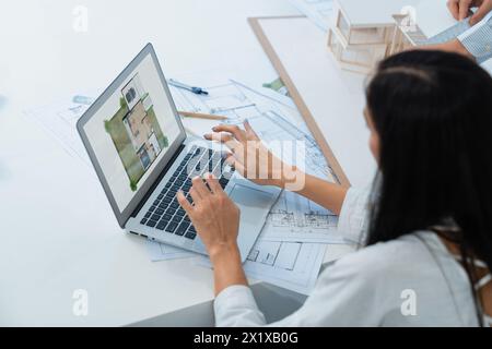 Wunderschöner kaukasischer Architekt mit Laptop, während ein intelligenter Kollege das Hausmodell mit Bauplan misst, Ausrüstung auf dem Tisch platziert. Kreatives Geschäft Stockfoto