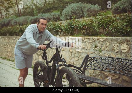 Europäischer junger Mann, der sein Elektro-Mountainbike die Straße rauf schiebt. Aktiver Lebensstil. Sport. Abenteuerfahrt mit dem Fahrrad. Verwendung von El-Bike ad ecologi Stockfoto