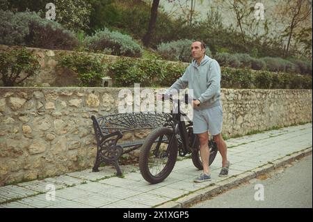 Im Freien ein Foto von einem glücklichen jungen sportlichen Mann, der sein elektrisches batteriebetriebenes Fahrrad, Mountainbike, die Straße hochschiebt. Aktiver Lebensstil. Sport. Erw Stockfoto