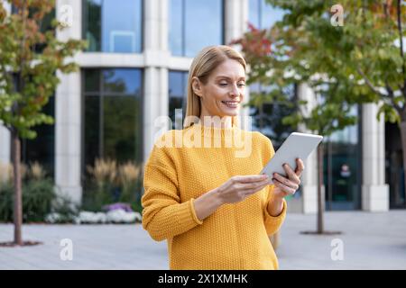 Eine professionelle Geschäftsfrau in einem gelben Pullover verwendet ein digitales Tablet außerhalb eines modernen Bürogebäudes. Sie sieht zufrieden und konzentriert aus. Stockfoto