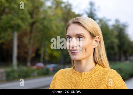 Eine fröhliche Frau mittleren Alters in einem hellgelben Pullover spaziert fröhlich in einem urbanen Park, umgeben von Herbstfarben. Stockfoto