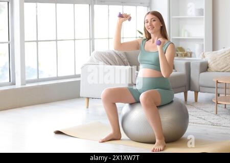 Sportliche junge schwangere Frau, die zu Hause mit Kurzhanteln am Fitball trainiert Stockfoto