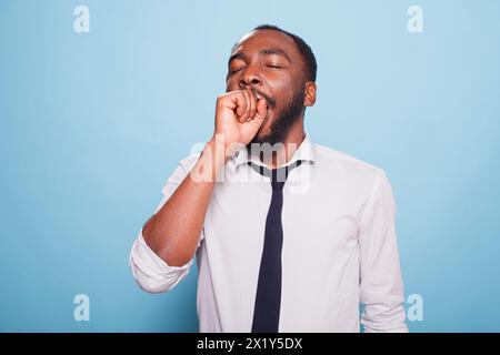 Müder schwarzer Mann gähnt mit seinem Mund bedeckt vor isoliertem Hintergrund. Erschöpfter afroamerikanischer Unternehmer, der in die Kamera schaut, sich müde und überarbeitet fühlt. Stockfoto