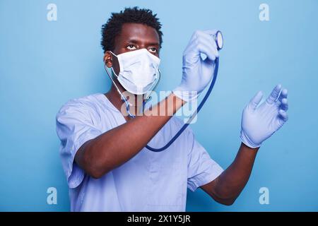 Männliche Pflegefachkraft hält Stethoskop, um Herzschlag von Kardiologiepatienten zu untersuchen. Afroamerikaner mit Handschuhen und Gesichtsmaske als Kardiologe bekämpft Coronavirus-Epidemie. Stockfoto