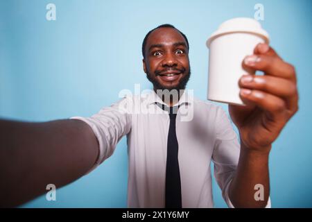 Schwarzer Mann, der einen Videoanruf hat, während er eine Tasse Kaffee vor isoliertem Hintergrund hält. Ein lächelnder afroamerikaner in weißem Hemd macht ein Selfie-Foto mit einem Getränkebehälter in der Hand. Stockfoto