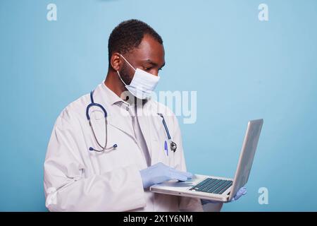 Arzt mit Latexhandschuhen und Gesichtsmaske, die einen Laptop hält, um Befunde einer klinischen Diagnose zu überprüfen. Der tragbare Computer wird von einem Mediziner gehalten, der Laborkittel und Stethoskop trägt. Stockfoto