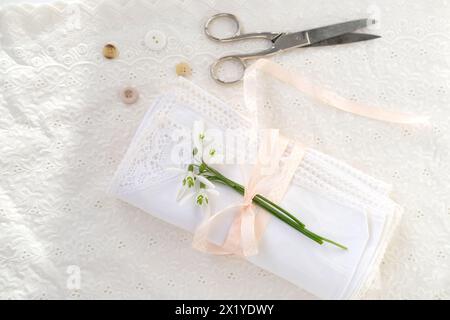 Weiße Leinenserviette, mit handgemachter Spitze beschnitten, in mehreren Schichten gerollt, zarte Schneeglöckchenblume, Schere, Knöpfe, Band ist auf Lichttisch, Conce Stockfoto