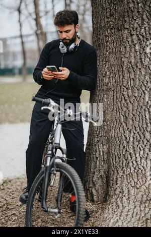 Ein aktiver Mann macht eine Pause gegen einen Baum im Park, um sein Smartphone mit Kopfhörern um den Hals und einem Fahrrad an seiner Seite zu überprüfen. Stockfoto