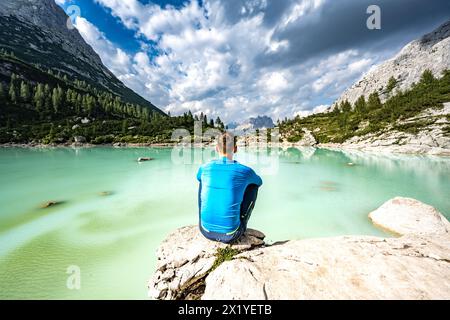 Beschreibung: Der Mann sitzt auf einem großen Felsen am türkisfarbenen Sorapis-See und genießt am Nachmittag die Aussicht auf die beeindruckende Cadini di Misurina. Lake Sorapis, Dolom Stockfoto