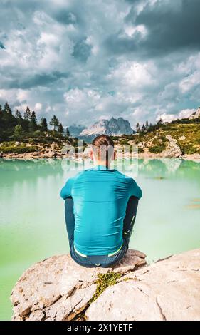 Beschreibung: Der Mann sitzt auf einem großen Felsen am türkisfarbenen Sorapis-See und genießt am Nachmittag die Aussicht auf die beeindruckende Cadini di Misurina. Lake Sorapis, Dolom Stockfoto