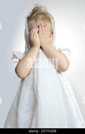 Kleines Kind, ein schüchternes Mädchen in einem weißen Kleid bedeckte ihr Gesicht mit ihren Handflächen aus Peinlichkeit, das Konzept von Kindheit, Elternschaft, Babyweinen von r Stockfoto