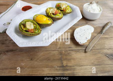 Gebackene Avocado gefüllt mit Ei, Gemüse, auf einer weißen Schale liegen, Brot mit Quark beschmiert, Leinenserviette mit Stickerei, Konzept der Post-Holiday d Stockfoto