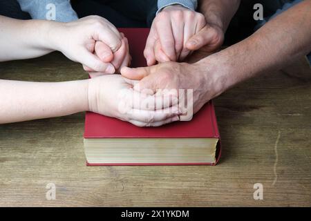 Nahaufnahme der Hände von zwei Männern und einer Frau gefaltet in einer Gebetsgeste auf einer dicken Familienbibel in einem dunkelroten Cover, das Konzept des ewigen Christen Stockfoto