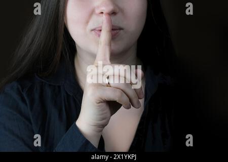 Nahaufnahme Frau, junges Mädchen in einer schwarzen Bluse mit langen Haaren drückte ihren Finger an ihre Lippen, regt zur Stille, Geste auf dunklem Hintergrund, Verhüllung an Stockfoto