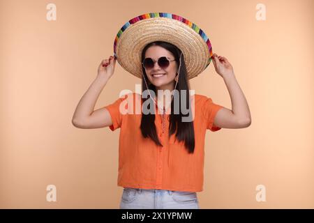 Junge Frau mit mexikanischem Sombrero-Hut und Sonnenbrille auf beigefarbenem Hintergrund Stockfoto