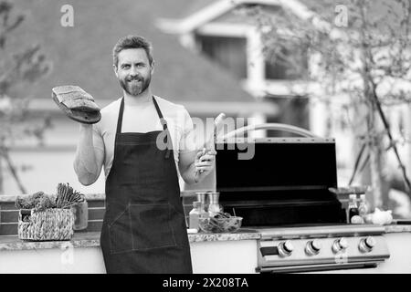 Glücklicher gegrillter Lachsfisch bei man trägt die Schürze des Küchenchefs. Foto des Küchenchefs mit gegrilltem Lachs. Koch mit gegrilltem Lachs. Küchenchef kocht gegrillten Lachs im Freien Stockfoto