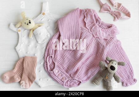 Set mit pinkfarbenen Kleidern und Accessoires für Neugeborene. Gestricktes Spielzeug Hase und Hund, Strampler, Socken und Handband auf weißem Holzhintergrund. Babydusche Stockfoto