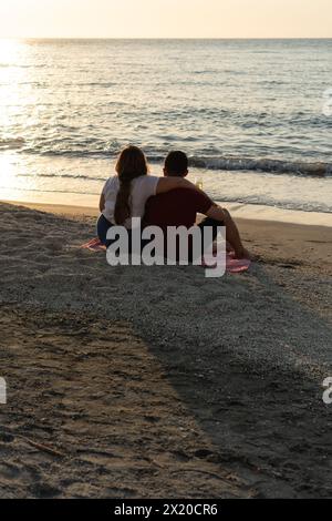 Rückansicht eines Paares in einer Umarmung, das die Wellen bei Sonnenuntergang beobachtet. Stockfoto