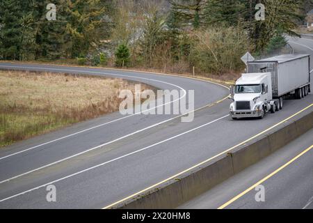 Weißer, großer Sattelzugmaschine mit flachem Fahrerhaus für den Transport von Gütern in einem überdachten Auflieger auf der geteilten Highwa Stockfoto