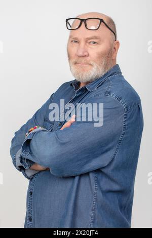 Porträt eines attraktiven grauhaarigen Mannes mit Brille, der ein Jeanshemd auf grauem Hintergrund trägt. Bärtiger Großvater europäischer Art. Stockfoto