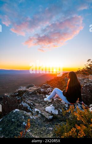 Frau, die am Rande der Reed Lookout Klippe sitzt und den Sonnenuntergang über Victoria Valley in den Grampians Mountains in Victoria, Australien, genießt Stockfoto