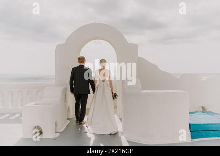 Eine Braut und ein Bräutigam spazieren an einem sonnigen Tag durch einen weißen Torbogen. Die Braut trägt ein weißes Kleid und der Bräutigam einen Anzug. Der Torbogen Stockfoto