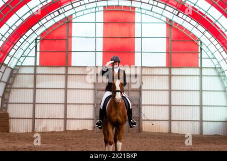 Reiterinnen grüßen in roter Halle. Reittraining für Frauen in einer roten Arena. Dressur. Weiblicher Jockey im Uniform-Reitpferd. Eques Stockfoto