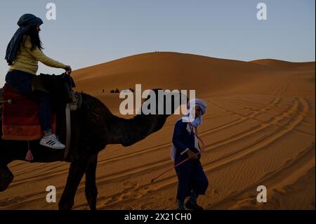 Marokkanischer Kameleer in Turban und Gewand hält das Zaumzeug eines Kamels, mit einem Touristen an Bord, vor dem Hintergrund einer Wüstendüne von Merzouga. Stockfoto