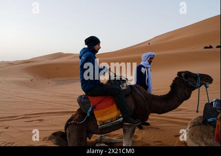 Ein Mann mittleren Alters bereitet sich darauf vor, ein Kamel abzusteigen, unterstützt von einem lokalen Handler, während das Tier kniet, mit einer aufsteigenden Sanddünen-Kulisse in der marokkanischen Wüste. Stockfoto