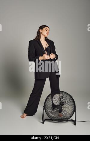 Eine stilvolle Frau in Übergröße strahlt Vertrauen in einen maßgeschneiderten Anzug aus, der anmutig neben einem drehenden Fan vor grauem Hintergrund steht. Stockfoto