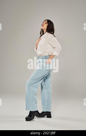 Eine Frau in Übergröße strahlt Vertrauen aus in einem weißen Hemd und einer blauen Jeans vor grauem Hintergrund. Stockfoto